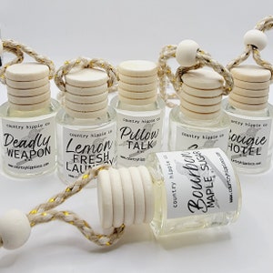 May include: Six small glass bottles with wooden tops and string hangers. Each bottle has a white label with black text and a small bird graphic. The labels read "Deadly Weapon", "Lemon Fresh Laundry", "Pillow Talk", "Bourbon Maple Sugar", and "Soughie Hotel".