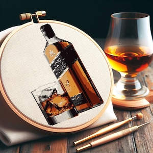 May include: A close-up of a cross-stitch embroidery hoop with a brown bottle and a glass of whiskey. The bottle has a black label with white text. The glass is filled with ice and whiskey.