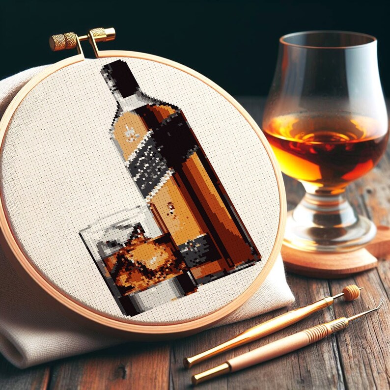 May include: A close-up of a cross-stitch embroidery hoop with a brown bottle and a glass of whiskey. The bottle has a black label with white text. The glass is filled with ice and whiskey.