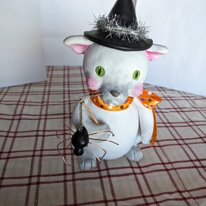 May include: White ceramic cat figurine wearing a black witch hat with silver tinsel trim, holding a black spider on a wire. The cat has green eyes and pink cheeks and is wearing a scarf with orange and black polka dots.