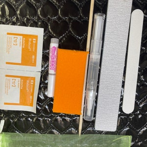 May include: A set of nail art supplies in a green mesh bag. The set includes a nail file, a nail buffer, a bottle of nail glue, a bottle of alcohol prep, and a wooden cuticle pusher.