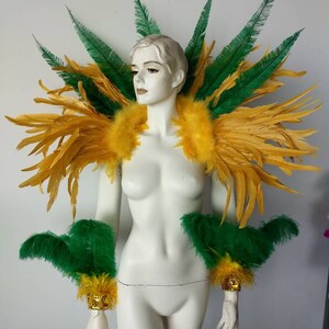 May include: A yellow and green feather costume shoulder piece with matching arm cuffs. The shoulder piece is large and dramatic, with a wide, rounded shape. The arm cuffs are smaller and more delicate, with a narrow, pointed shape.