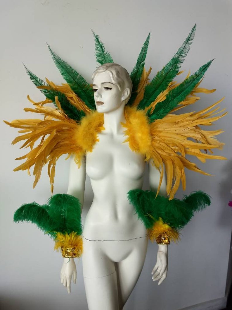 May include: A yellow and green feather costume shoulder piece with matching arm cuffs. The shoulder piece is large and dramatic, with a wide, rounded shape. The arm cuffs are smaller and more delicate, with a narrow, pointed shape.