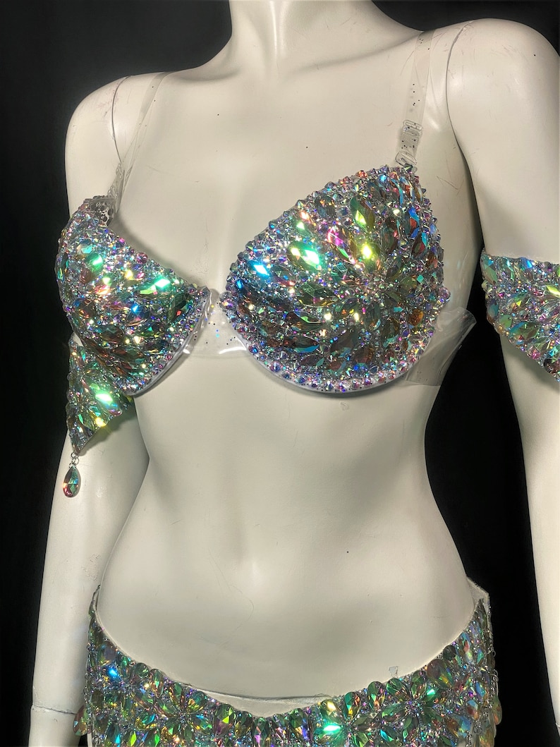 May include: A two-piece iridescent crystal costume set with a bra and a skirt. The bra has clear straps and the skirt has a high waist.
