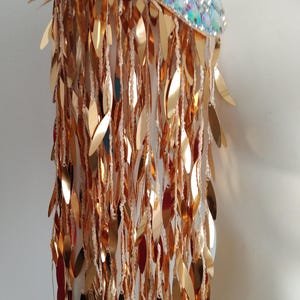 May include: A shoulder epaulet featuring a top panel covered in iridescent, multi-colored reflective stones. Attached to the base are numerous long, dangling strands of metallic gold, leaf-shaped sequins that create a cascading fringe effect.