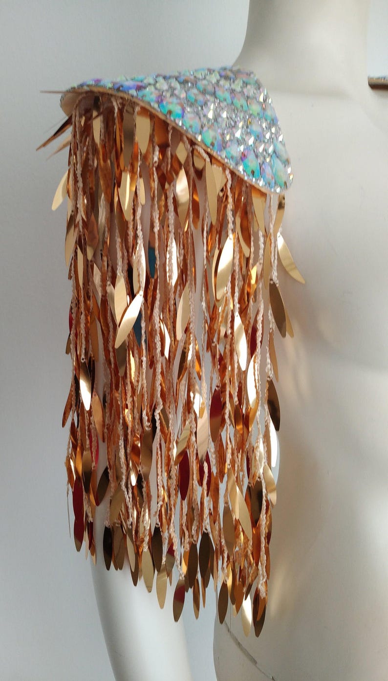 May include: A shoulder epaulet featuring a top panel covered in iridescent, multi-colored reflective stones. Attached to the base are numerous long, dangling strands of metallic gold, leaf-shaped sequins that create a cascading fringe effect.