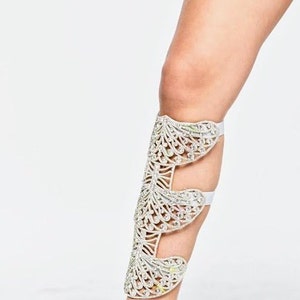 Puede incluir: Una pierna blanca con una bota de tac&oacute;n abierta de color plateado adornada con cristales. La bota tiene un dise&ntilde;o delicado e intrincado con m&uacute;ltiples capas de cristales.