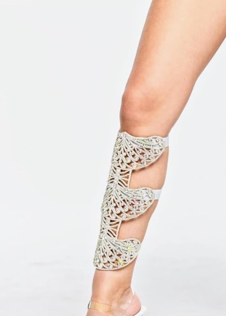 Puede incluir: Una pierna blanca con una bota de tac&oacute;n abierta de color plateado adornada con cristales. La bota tiene un dise&ntilde;o delicado e intrincado con m&uacute;ltiples capas de cristales.