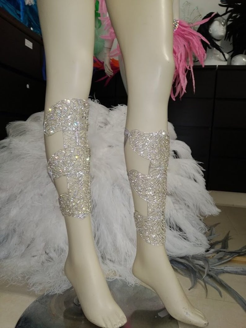 Puede incluir: Un par de brazaletes de pierna plateados con un dise&ntilde;o delicado e intrincado. Los brazaletes est&aacute;n adornados con brillantes pedrer&iacute;a y se llevan en las piernas de un maniqu&iacute;.