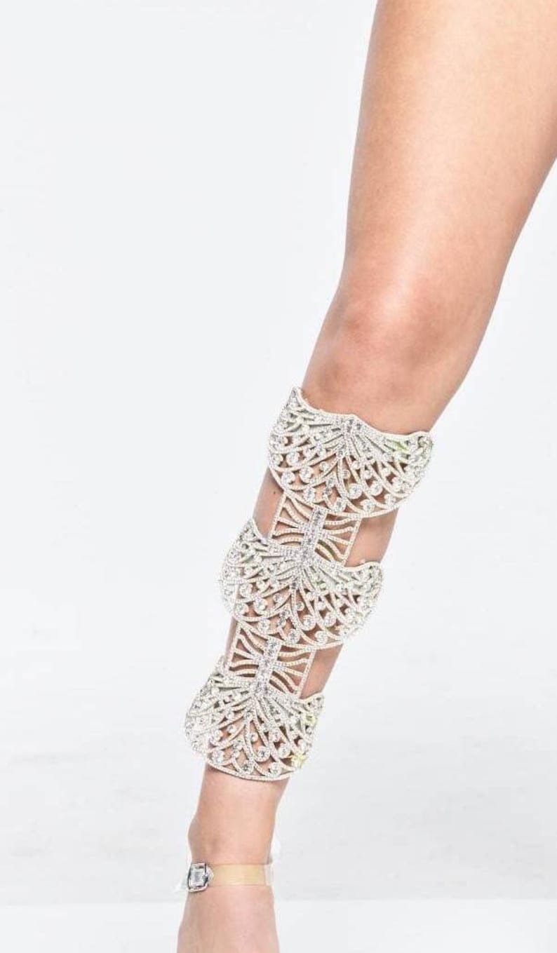 Puede incluir: Un brazalete de pierna plateado con un dise&ntilde;o delicado e intrincado que presenta m&uacute;ltiples capas de adornos de cristal. El brazalete se lleva en la pierna de una persona, extendi&eacute;ndose desde el tobillo hasta la rodilla.