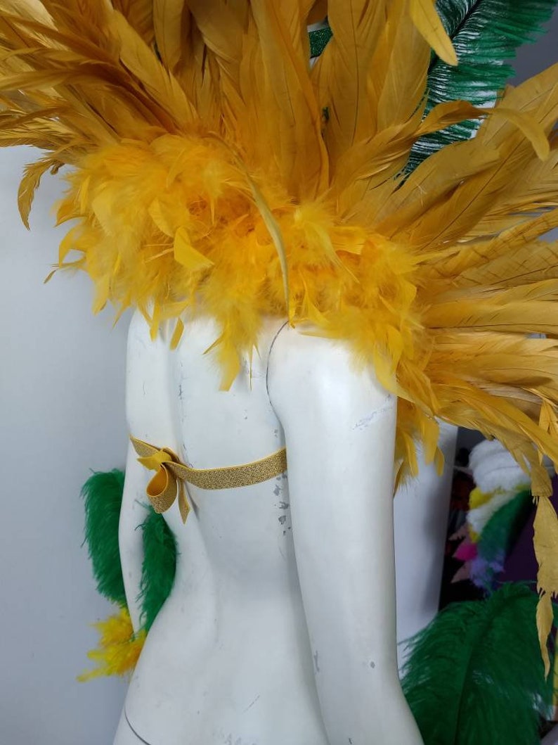 May include: A yellow feather costume piece with a gold ribbon. The feather piece is worn on the shoulders and is designed for a costume or performance.