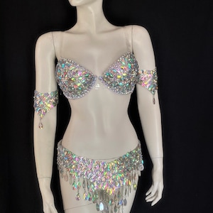 May include: A silver bra and belt set with iridescent rhinestones. The bra has a clear strap and the belt has a fringe of dangling rhinestones.