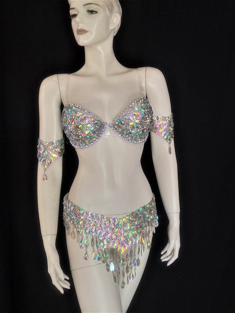 May include: A silver bra and belt set with iridescent rhinestones. The bra has a clear strap and the belt has a fringe of dangling rhinestones.