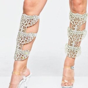 Puede incluir: Un par de polainas blancas con un dise&ntilde;o decorativo de mariposa hecho de pedrer&iacute;a. Las polainas se llevan en las piernas de una persona y se extienden desde el tobillo hasta la rodilla.