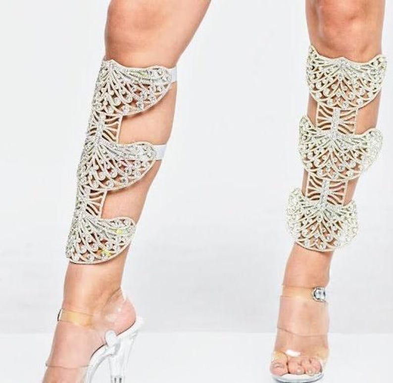 Puede incluir: Un par de polainas blancas con un dise&ntilde;o decorativo de mariposa hecho de pedrer&iacute;a. Las polainas se llevan en las piernas de una persona y se extienden desde el tobillo hasta la rodilla.