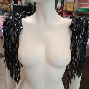 May include: A pair of black shoulder epaulets featuring dense, dangling sequin fringe. The top sections are embellished with clusters of round, reflective black sequins and small clear crystals, creating a textured, shimmering effect on the shoulders.