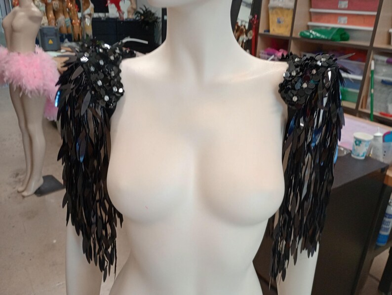 May include: A pair of black shoulder epaulets featuring dense, dangling sequin fringe. The top sections are embellished with clusters of round, reflective black sequins and small clear crystals, creating a textured, shimmering effect on the shoulders.