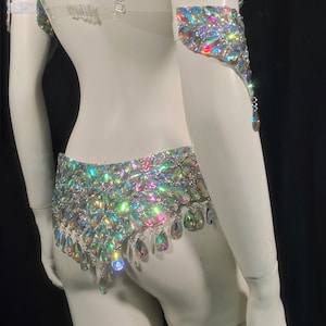 May include: A white costume set with iridescent crystal embellishments. The set includes a bra top, a belt, and arm cuffs.
