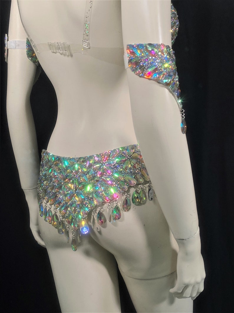 May include: A white costume set with iridescent crystal embellishments. The set includes a bra top, a belt, and arm cuffs.
