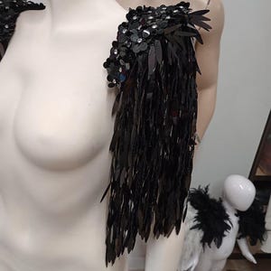 May include: A black shoulder epaulet featuring dense layers of shiny, elongated sequins and small floral-shaped embellishments with clear crystals. The piece is designed to drape over the shoulder, creating a fringe effect with reflective black material.