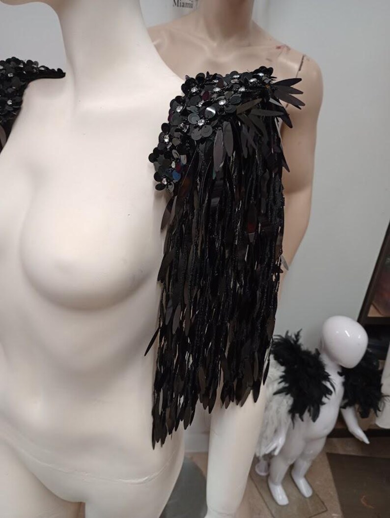 May include: A black shoulder epaulet featuring dense layers of shiny, elongated sequins and small floral-shaped embellishments with clear crystals. The piece is designed to drape over the shoulder, creating a fringe effect with reflective black material.