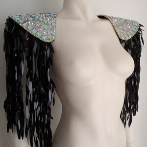 May include: A pair of iridescent crystal shoulder pieces with black fringe. The shoulder pieces are made of a fabric base covered in crystals and have a pointed shape. The fringe is made of black feathers and hangs down from the bottom of the shoulder pieces.