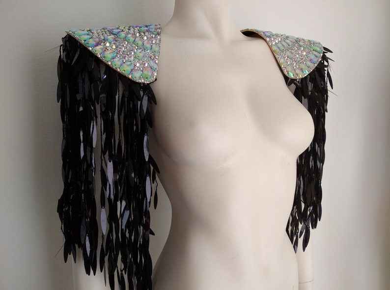 May include: A pair of iridescent crystal shoulder pieces with black fringe. The shoulder pieces are made of a fabric base covered in crystals and have a pointed shape. The fringe is made of black feathers and hangs down from the bottom of the shoulder pieces.