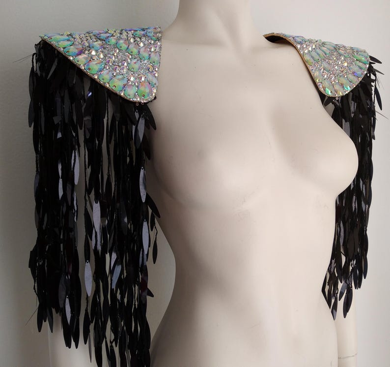 May include: A pair of shoulder epaulets featuring iridescent, multi-faceted crystals on the top panels. Long, dangling black sequin fringe hangs from the edges of each shoulder piece, creating a layered, textured effect across the upper arms.