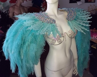 May include: A turquoise feather shoulder piece and matching bra top adorned with iridescent crystals. The garment features long, flowing turquoise feathers extending from the shoulders and a structured bra frame covered in sparkling, multi-colored rhinestones.
