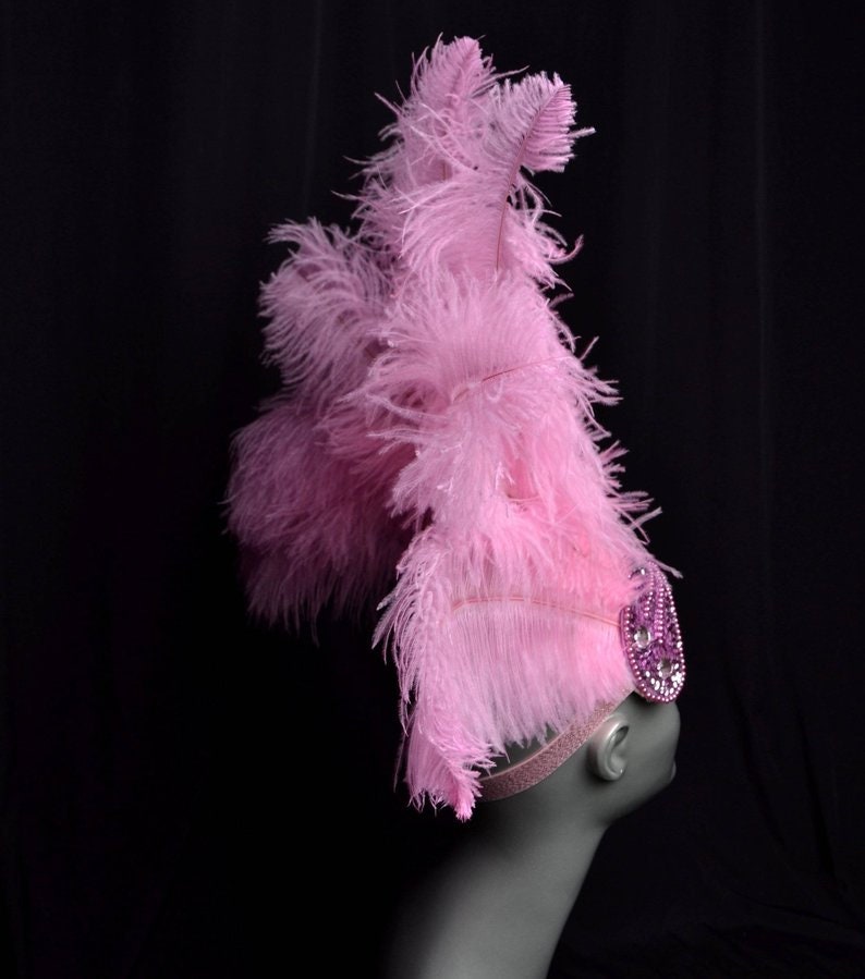 May include: A pink feather headpiece with a pink rhinestone embellishment. The headpiece is worn on a mannequin's head.