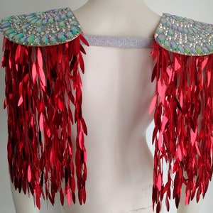 May include: A pair of shoulder epaulets featuring iridescent, multi-faceted crystals on the top panels and long, dangling red metallic sequin fringe. The two shoulder pieces are connected by a wide, shimmering silver elastic band.
