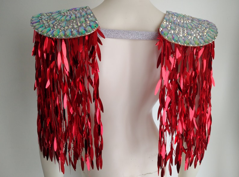 May include: A pair of shoulder epaulets featuring iridescent, multi-faceted crystals on the top panels and long, dangling red metallic sequin fringe. The two shoulder pieces are connected by a wide, shimmering silver elastic band.