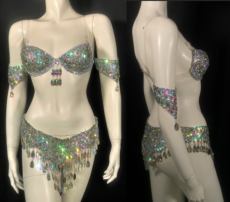 May include: A silver and iridescent belly dancing costume with a bra, belt, and arm cuffs. The costume is covered in rhinestones and has a fringe detail.