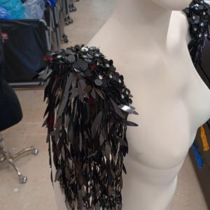 May include: A black shoulder epaulet featuring dense layers of dangling, leaf-shaped sequins and round, flat sequins. The piece is constructed with a structured base that sits atop the shoulder, creating a cascading fringe effect of dark, reflective material.