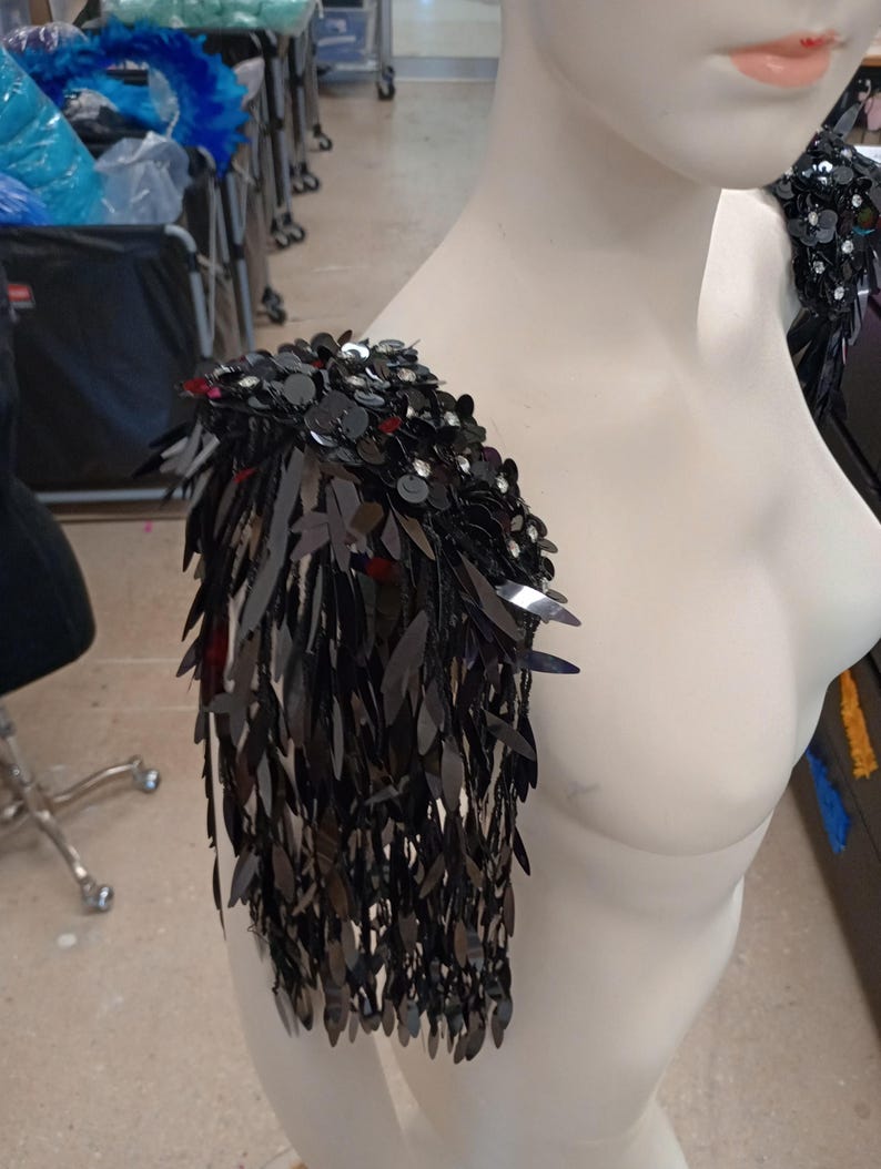 May include: A black shoulder epaulet featuring dense layers of dangling, leaf-shaped sequins and round, flat sequins. The piece is constructed with a structured base that sits atop the shoulder, creating a cascading fringe effect of dark, reflective material.