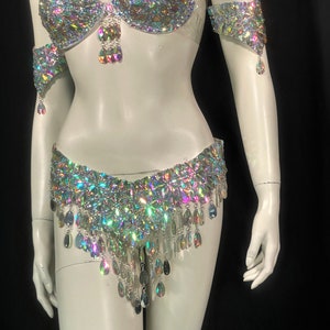 May include: A silver and iridescent costume set with a bra, belt, and arm cuffs. The set is covered in rhinestones and has dangling teardrop-shaped crystals.