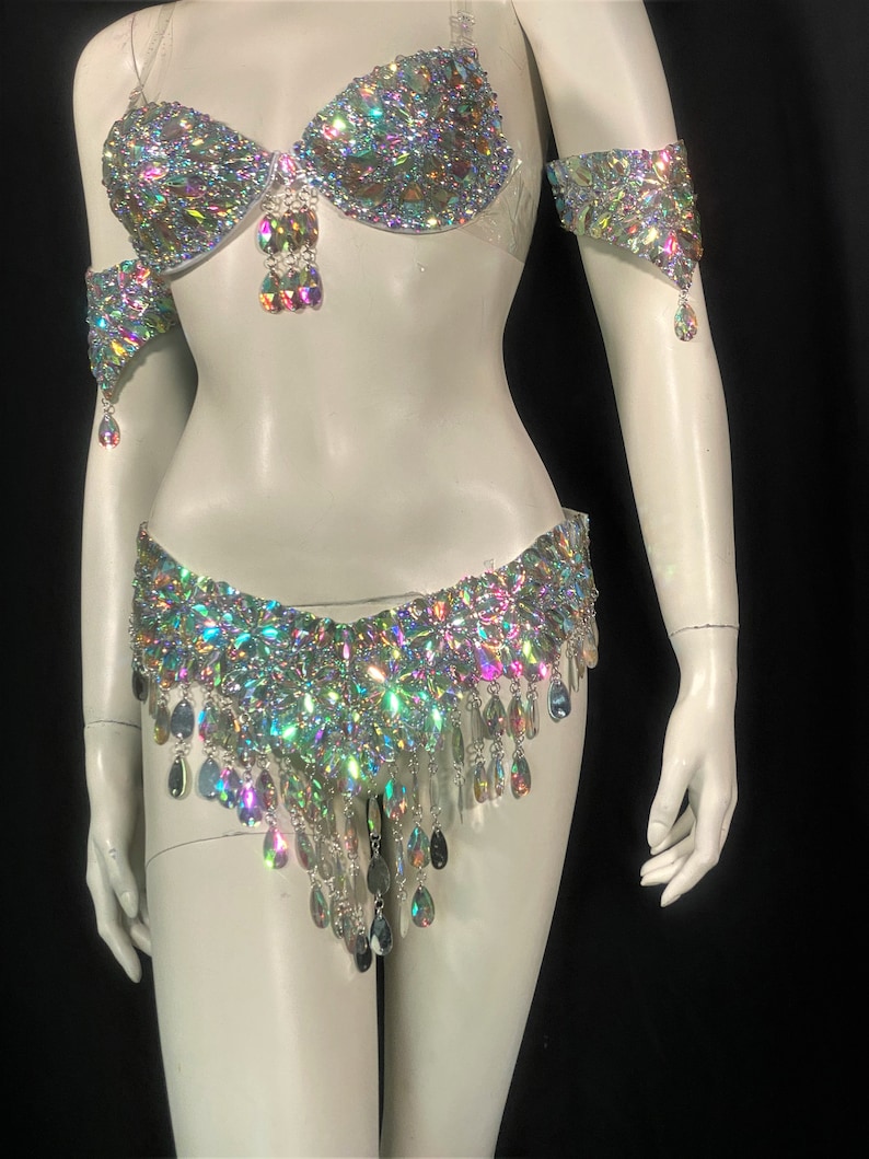 May include: A silver and iridescent costume set with a bra, belt, and arm cuffs. The set is covered in rhinestones and has dangling teardrop-shaped crystals.