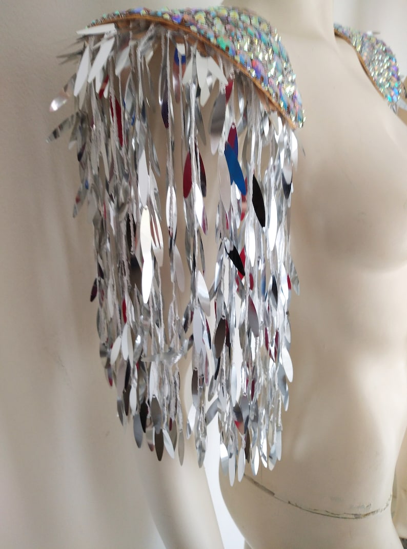 May include: A pair of shoulder epaulets featuring a top layer of iridescent, multi-colored sequins and a cascading fringe of long, reflective silver leaf-shaped metallic pieces that hang down to create a shimmering, high-contrast visual effect.