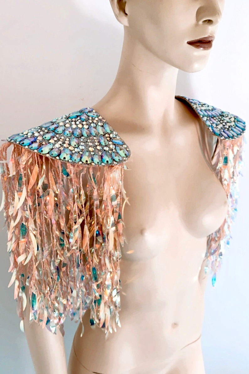 May include: A pair of shoulder epaulets featuring iridescent, multi-shaped crystals on the top panels. Long, shimmering, light-reflecting fringe strands in a pale peach hue hang from the edges, creating a cascading effect over the upper arms.