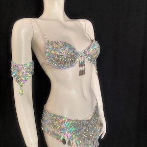 May include: A silver and iridescent costume set with a bra, belt, and arm cuff. The set is adorned with rhinestones and teardrop-shaped crystals.