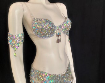Pode incluir: Um conjunto de fantasia de duas peças com um sutiã e um cinto de quadril adornados com cristais iridescentes multifacetados. O sutiã inclui alças transparentes e detalhes de cristais pendentes em forma de gota, com braçadeira a condizer.