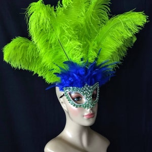 Pode incluir: Uma m&aacute;scara de carnaval com penas verdes e azuis e strass. A m&aacute;scara est&aacute; em uma cabe&ccedil;a de manequim.
