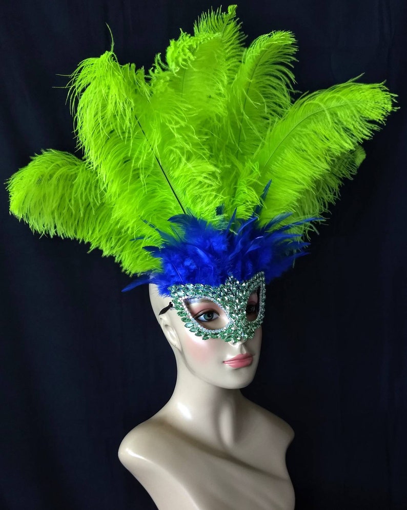 Pode incluir: Uma m&aacute;scara de carnaval com penas verdes e azuis e strass. A m&aacute;scara est&aacute; em uma cabe&ccedil;a de manequim.