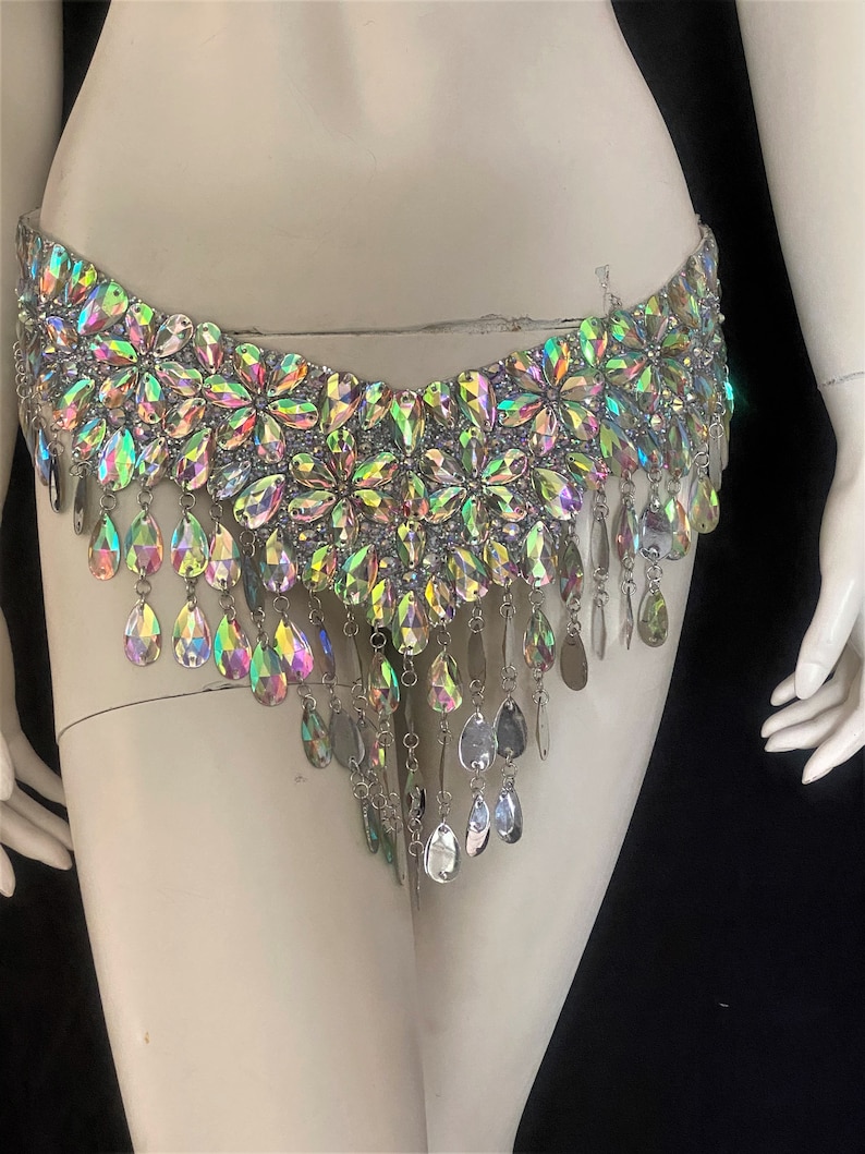 May include: A silver-toned belly chain with iridescent crystal embellishments. The chain features a row of flower-shaped crystals and a fringe of teardrop-shaped crystals.