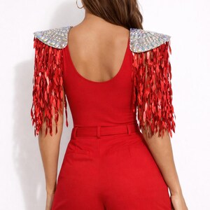 May include: A bright red bodysuit featuring shoulder epaulets adorned with iridescent crystals and long, dangling red sequin fringe. The garment includes a matching red high-waisted short with a thin belt, creating a cohesive monochromatic look.