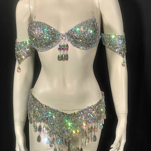 May include: A white belly dance costume with iridescent rhinestones and fringe. The costume includes a bra top, a belt, and arm cuffs.