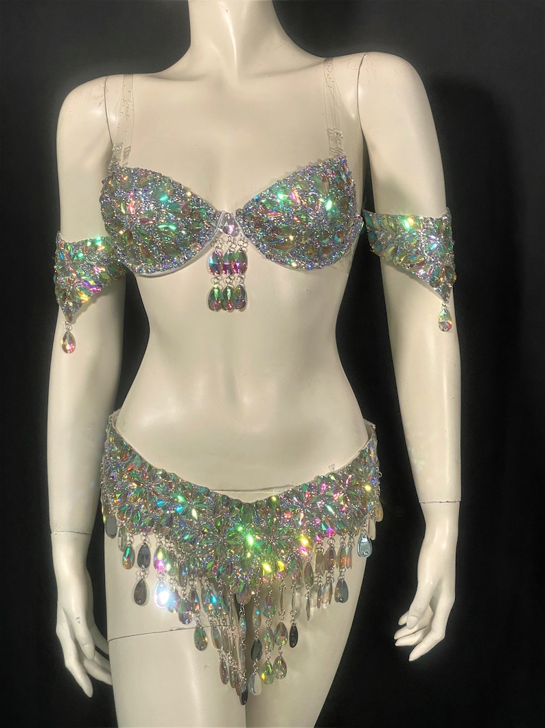 May include: A white belly dance costume with iridescent rhinestones and fringe. The costume includes a bra top, a belt, and arm cuffs.