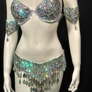 May include: A silver and iridescent costume set with a bra, belt, and arm cuffs. The bra and belt are covered in rhinestones and the belt has a fringe of teardrop-shaped crystals.