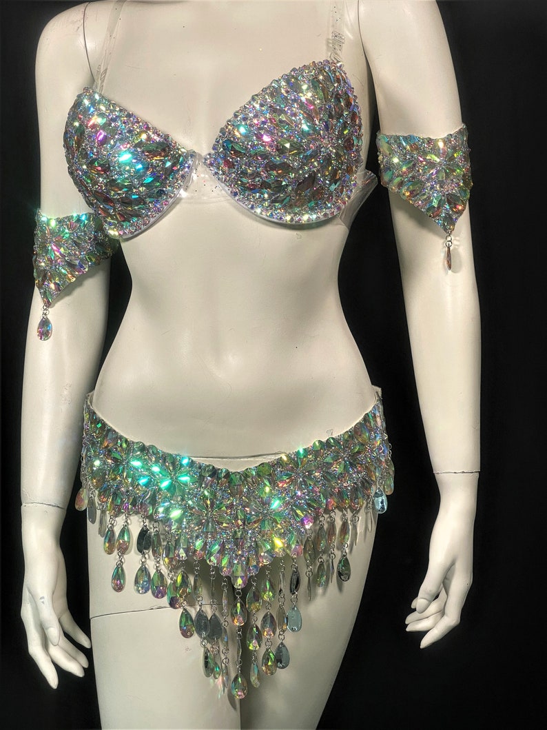 May include: A silver and iridescent costume set with a bra, belt, and arm cuffs. The bra and belt are covered in rhinestones and the belt has a fringe of teardrop-shaped crystals.