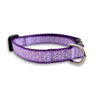 Puede incluir: Un collar para perro morado con un diseño estampado blanco. El collar tiene una hebilla plateada y un deslizador de plástico negro.